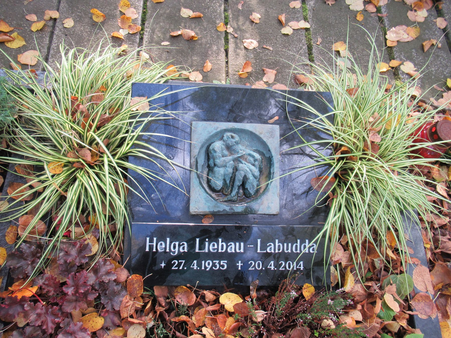 Dark recumbent headstone with a portrait relief among grasses and autumn leaves at Kaulsdorf Cemetery