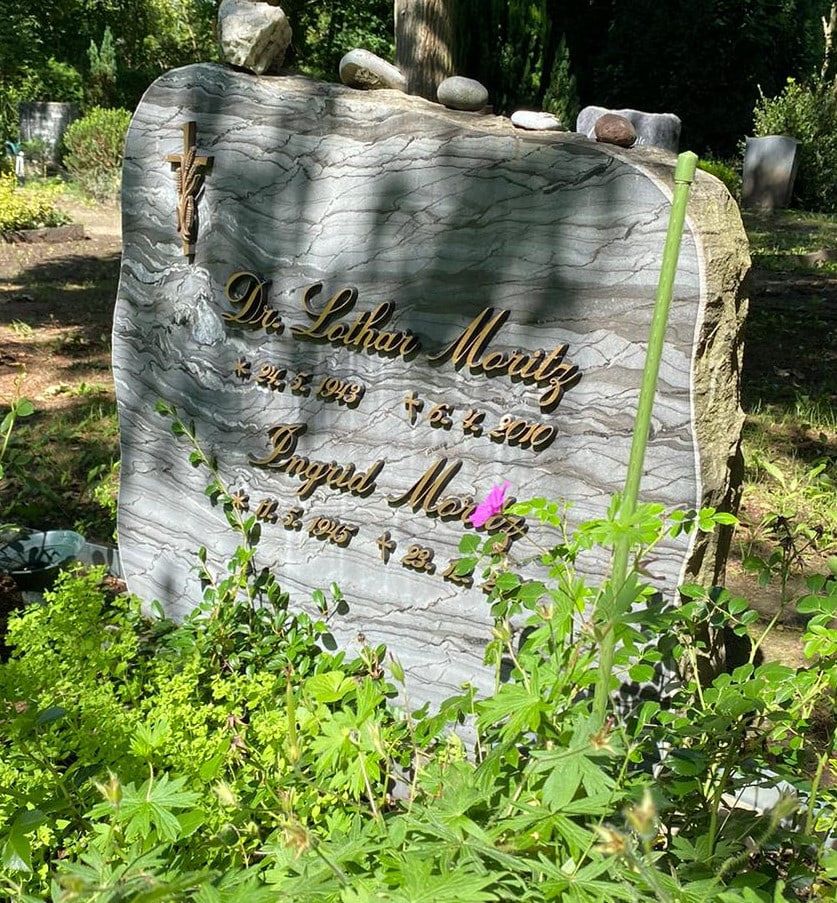 Light-coloured headstone with a curved top, planted border and decorative stones at Kaulsdorf Cemetery
